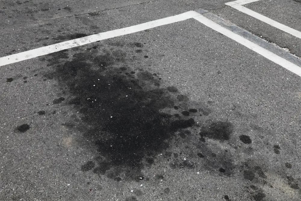 Dark motor oil stain soaked into asphalt beside painted parking stall markings