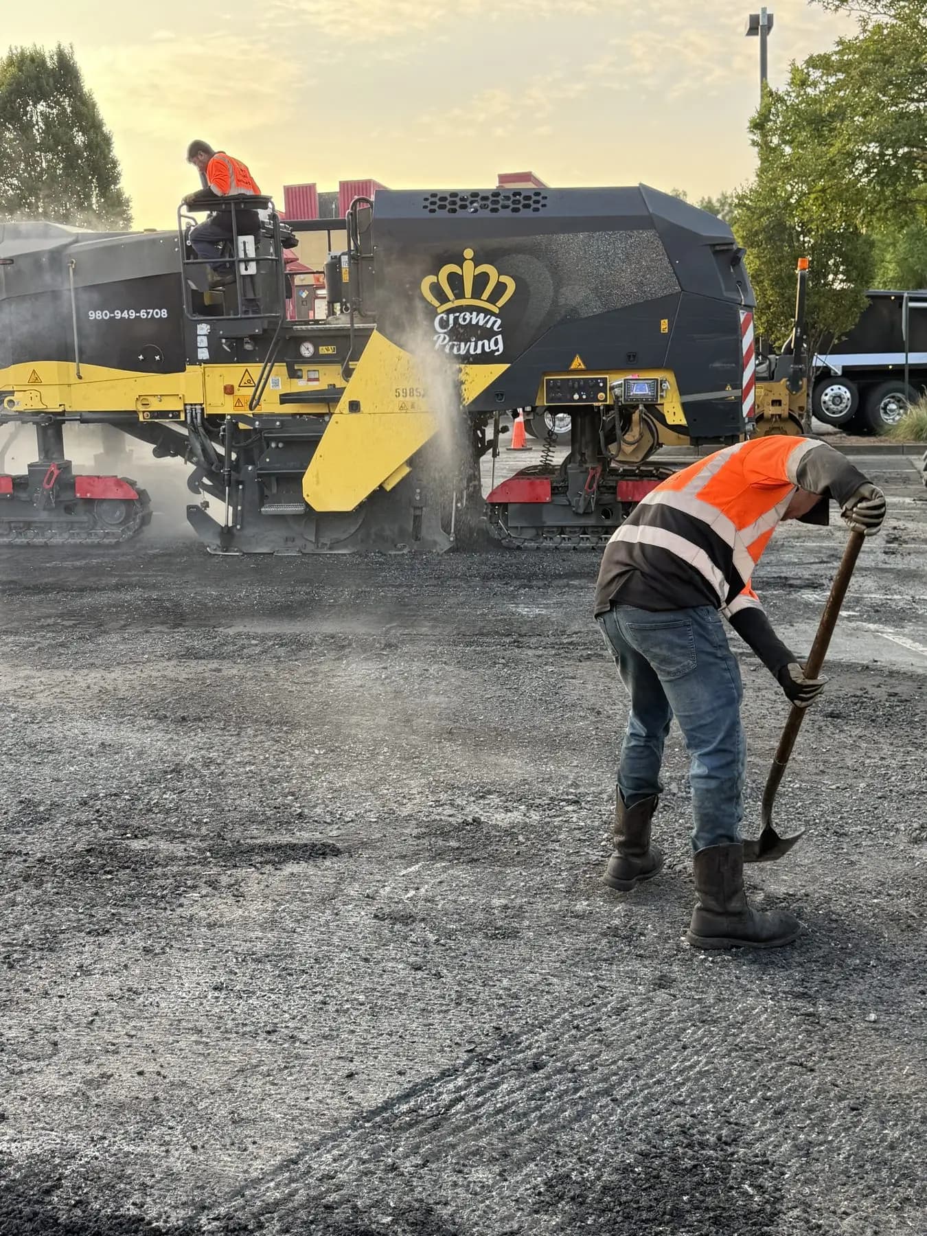 A Crown Paving milling machine cutting out old asphalt at first light, with a crew member clearing millings behind it