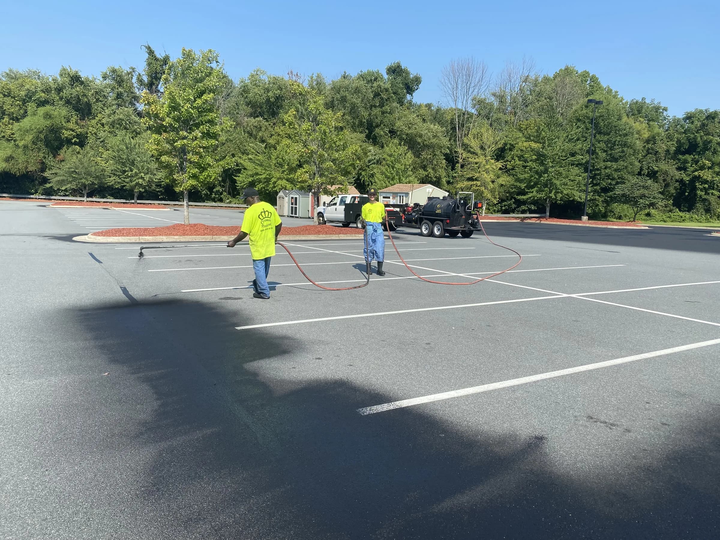 Two Crown Paving crew members spraying seal coat across a striped commercial lot, with the sealer tank behind them