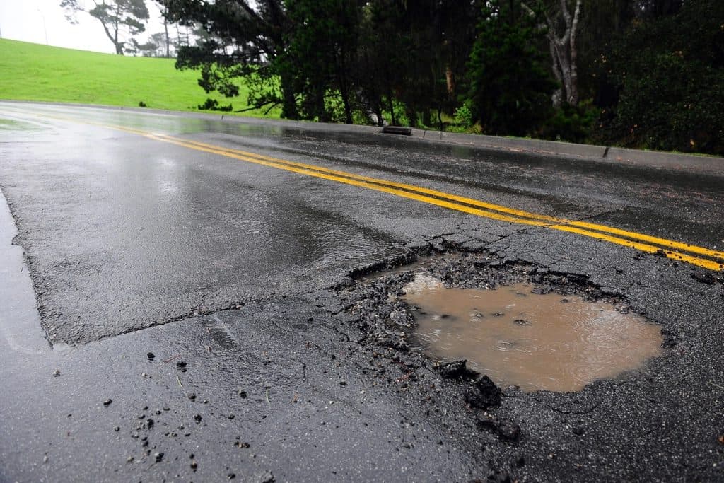 A pothole holding standing water in a wet asphalt surface, its broken edges crumbling beside an older patch